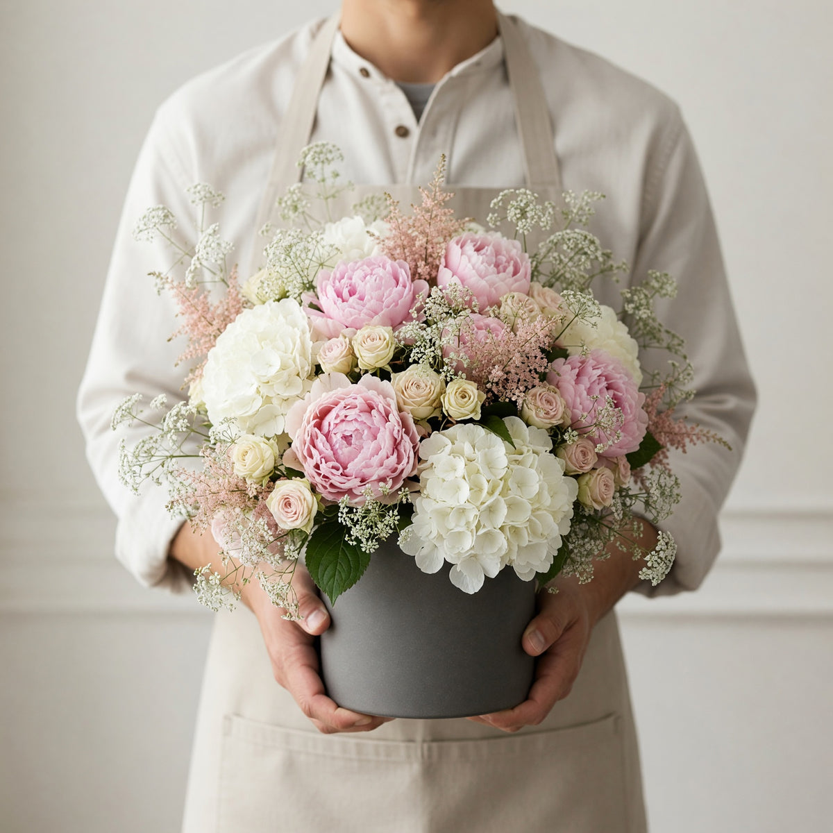 A person wearing a beige shirt and apron holds a large gray round box filled with a lush floral arrangement of pink and white flowers with green foliage, against a soft light gray background.