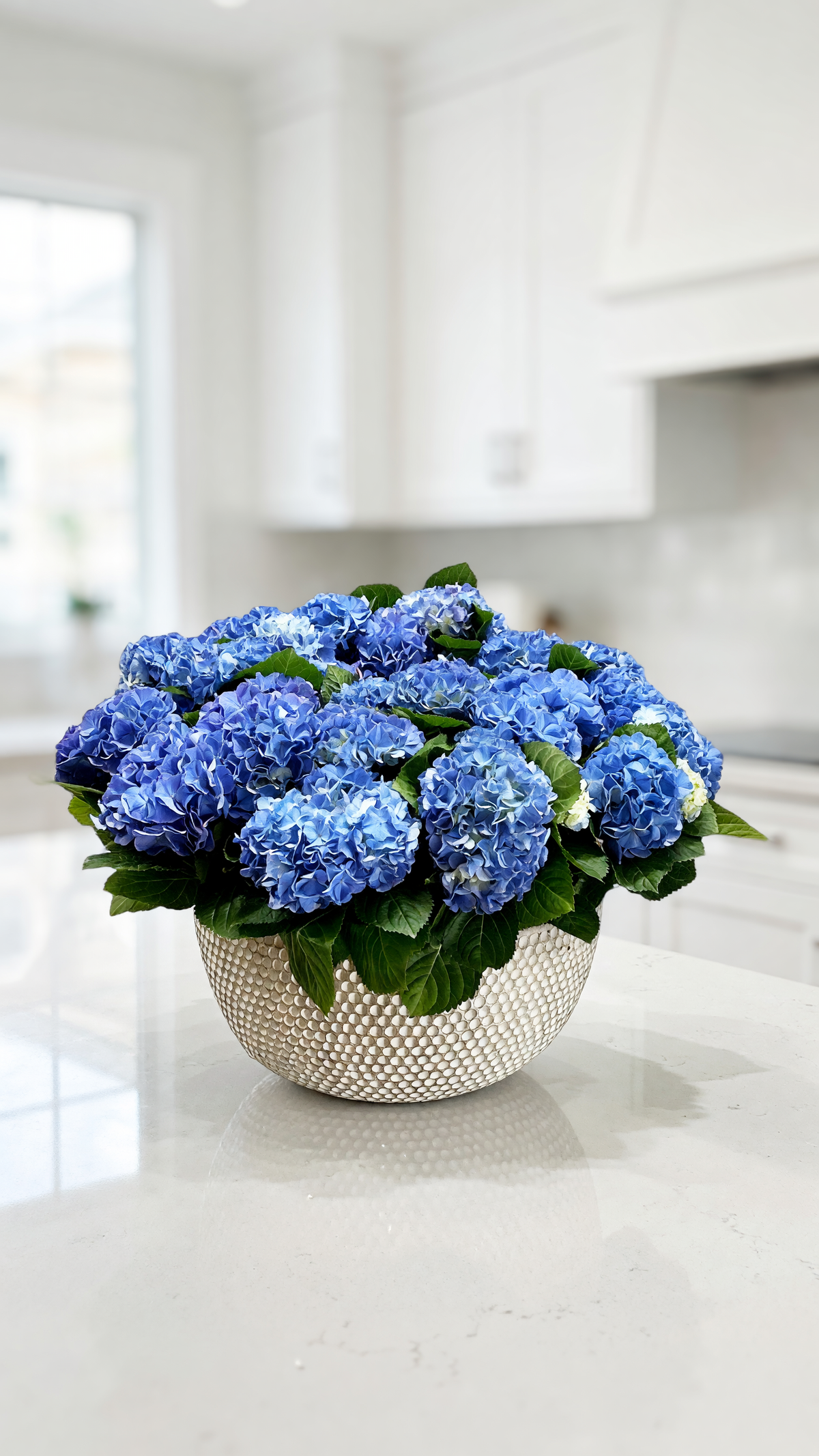 A vibrant bouquet of blue hydrangeas with green leaves arranged in a textured, metallic silver bowl, set on a white kitchen countertop with a blurred modern kitchen background.