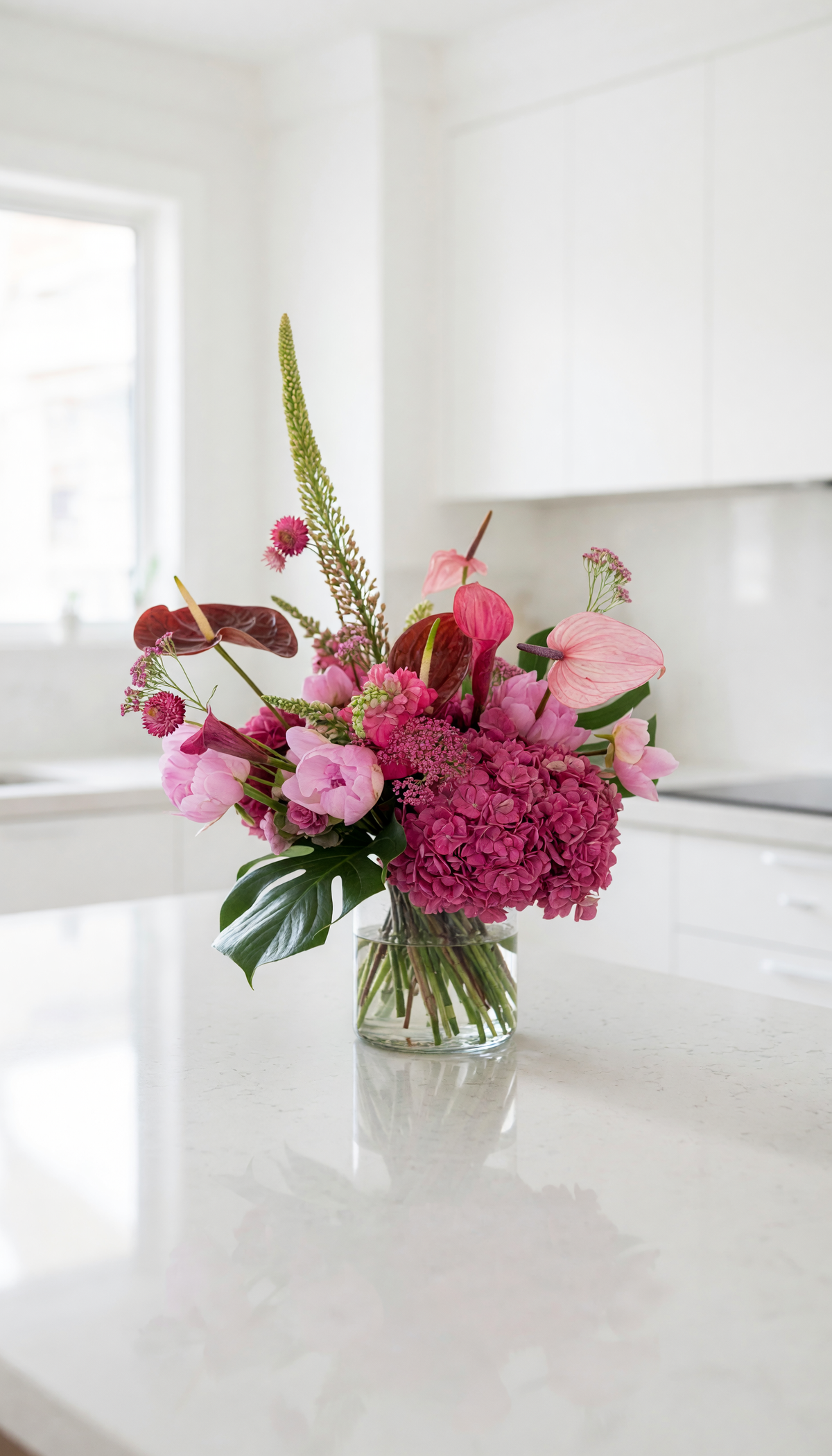 A vibrant pink and magenta floral arrangement in a clear glass jar vase, set on a white kitchen island with a blurred white kitchen background.