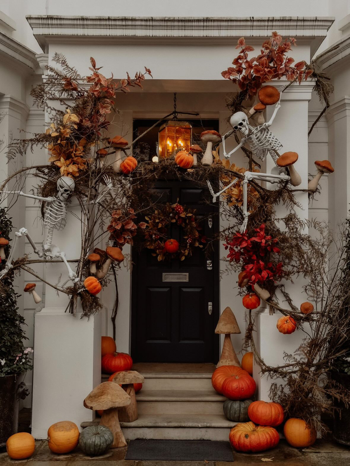 A Halloween-decorated front porch with a black door framed by skeletons, pumpkins, orange foliage, mushrooms, and a hanging lantern. The white house facade surrounds the entrance, with steps leading up to the door.