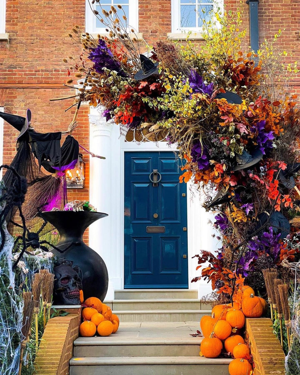 A festive Halloween front porch decorated with a large autumn floral arch, black witch hats, a cauldron, spider decor, and pumpkins on the steps in front of a brick house.