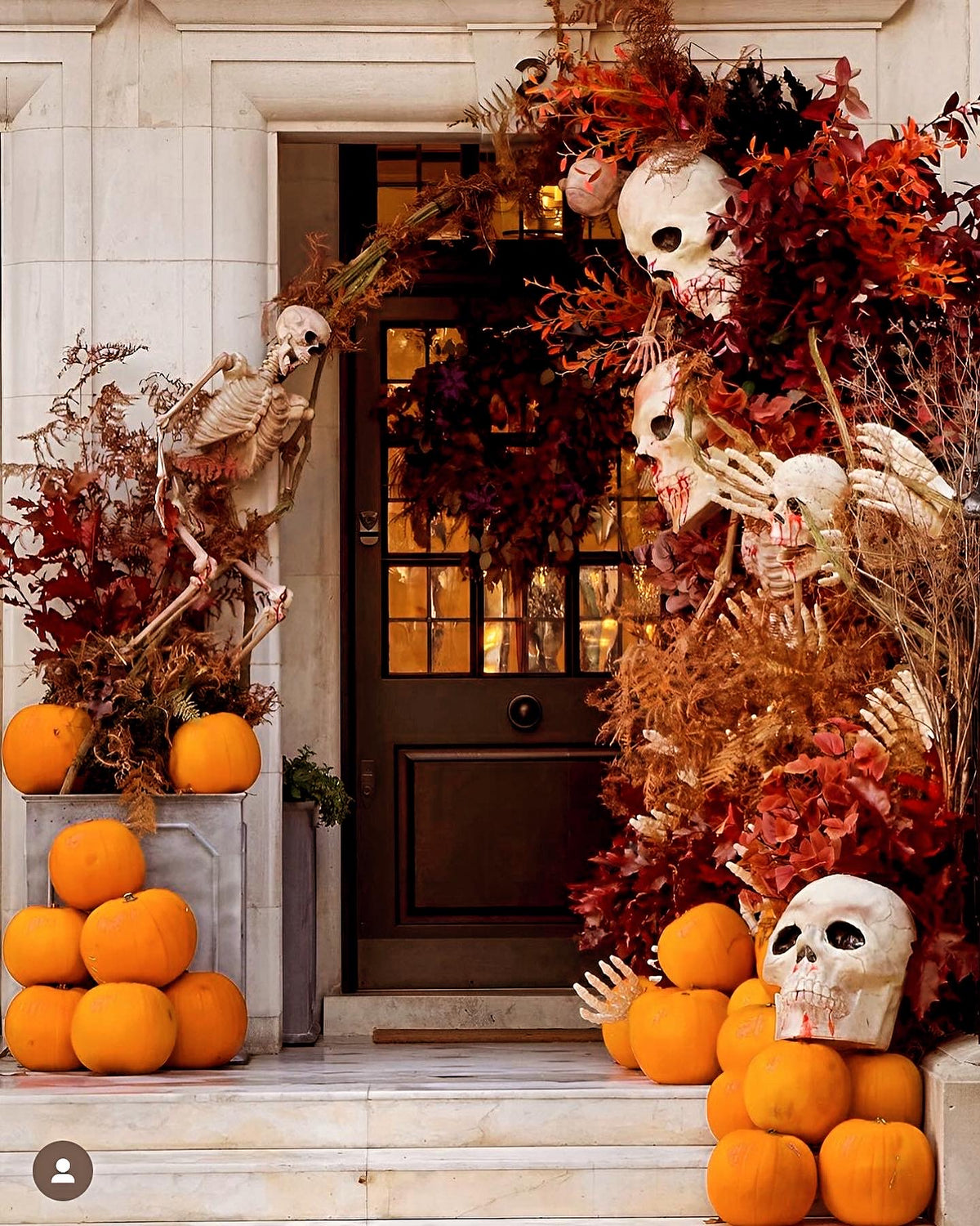 A Halloween front door decorated with a large arrangement of human skulls, dried autumn leaves, skeletons, pumpkins, and a wreath, set against a stone building entrance.