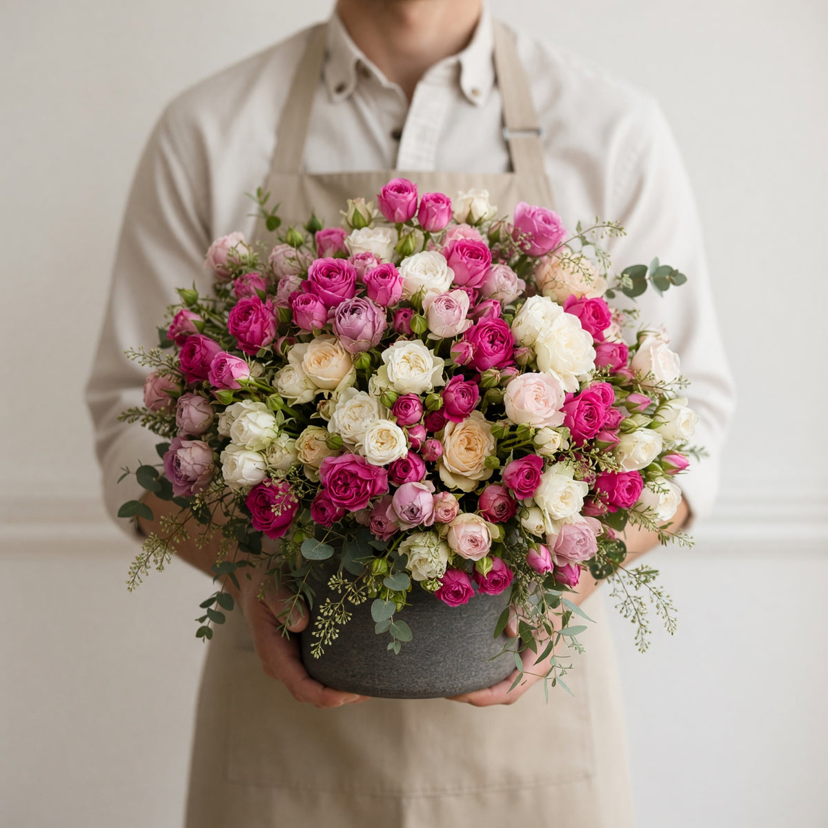 A person wearing a beige shirt and apron holds a large, round floral arrangement of pink, magenta, and white roses with green foliage in a gray pot against a light beige wall.