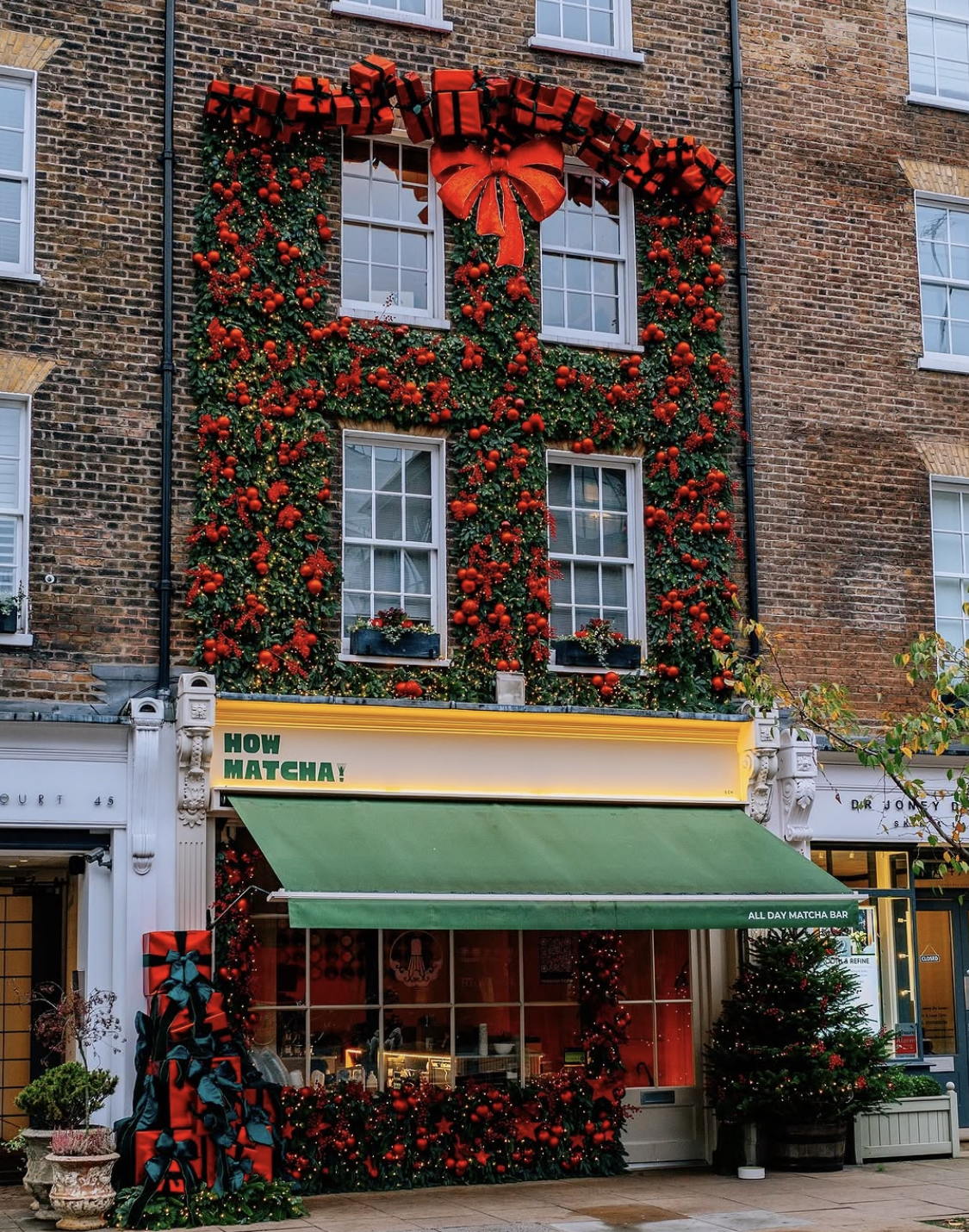 A brick building facade decorated for Christmas, with a green ivy-covered wall, large red bows, red ornaments, wrapped gifts, and a green awning above a storefront sign reading 'HOW MATCHA!'.