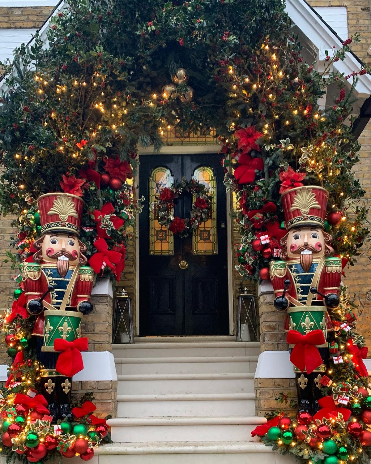 A front door decorated for Christmas with an arched garland of greenery and warm string lights, red bows, ornaments, and two large nutcracker figures flanking the steps.