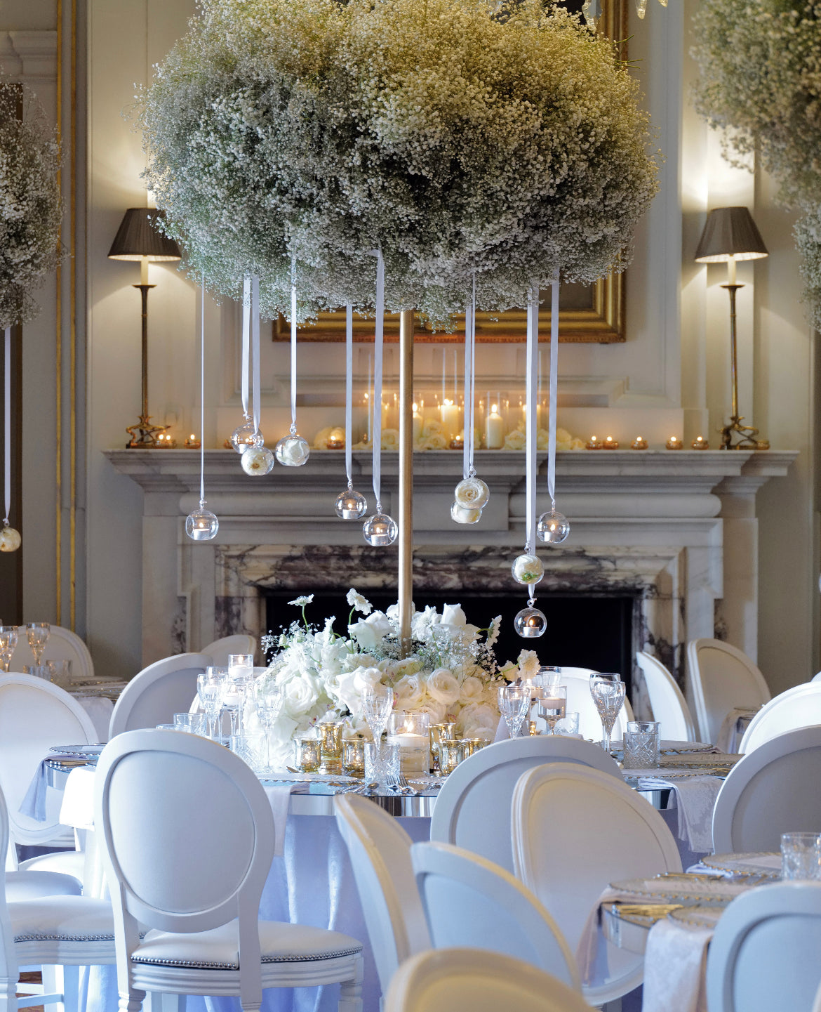 A large floral centerpiece hangs above a dining table in a formal indoor event space, with white chairs, lit candles, and a fireplace in the background.