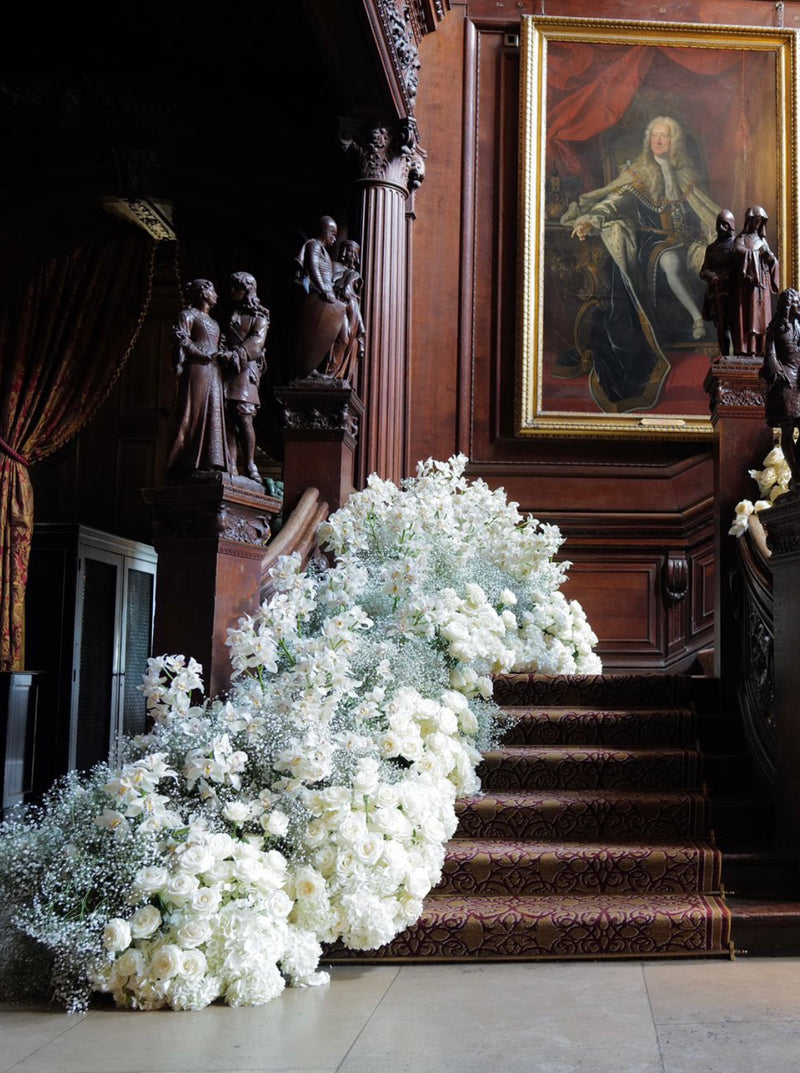 A large floral arrangement of white and pale green flowers cascades down an ornate dark wood staircase in a grand interior with statues and a large portrait painting on the wall.