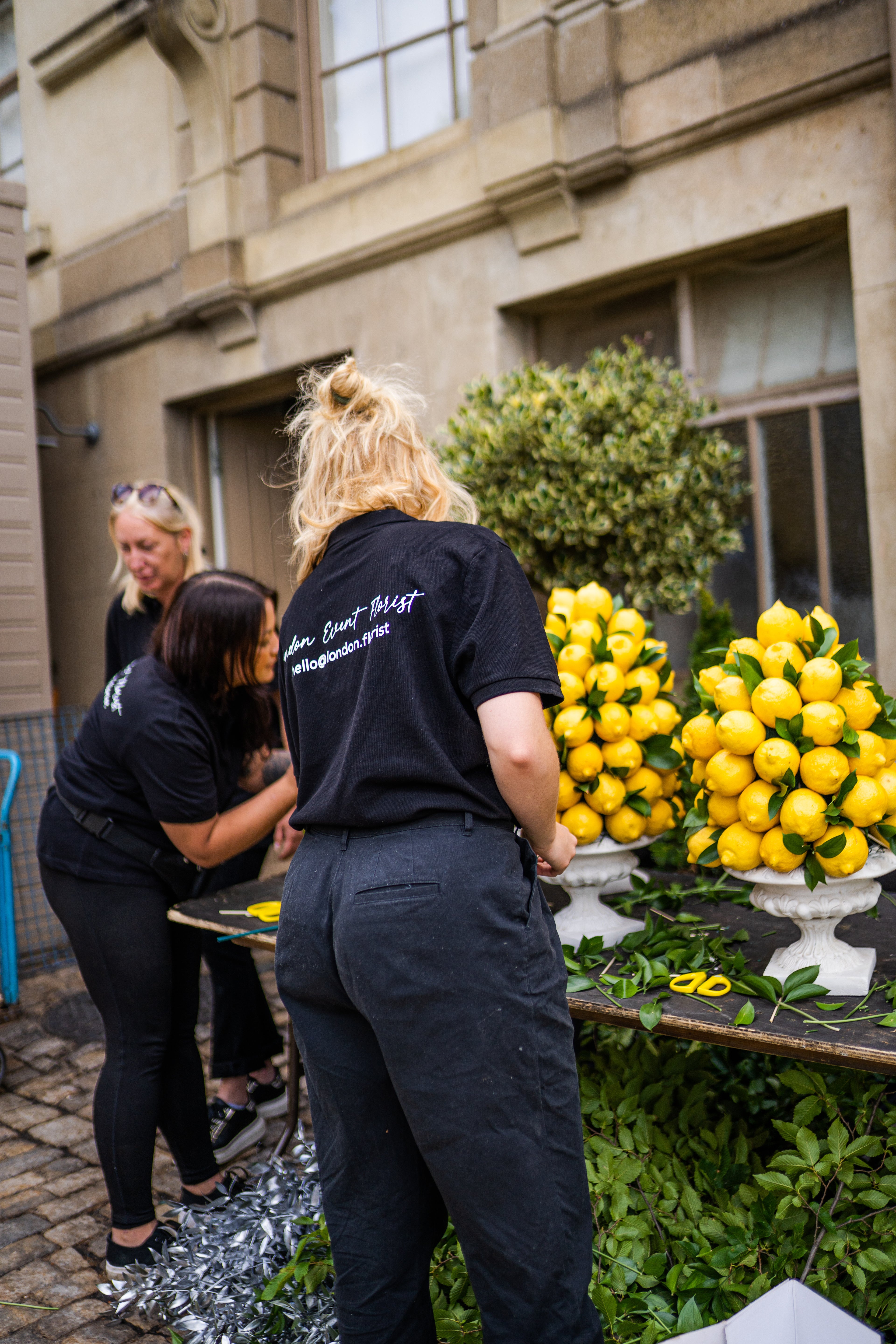 Three women wearing matching black t-shirts with white text on the back are gathered around a table displaying large clusters of fresh yellow lemons in white planters. The setting is outdoors in front of a stone building.