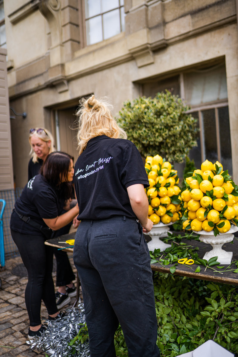 Three women wearing matching black t-shirts with white text on the back are gathered around a table displaying large clusters of fresh yellow lemons in white planters. The setting is outdoors in front of a stone building.