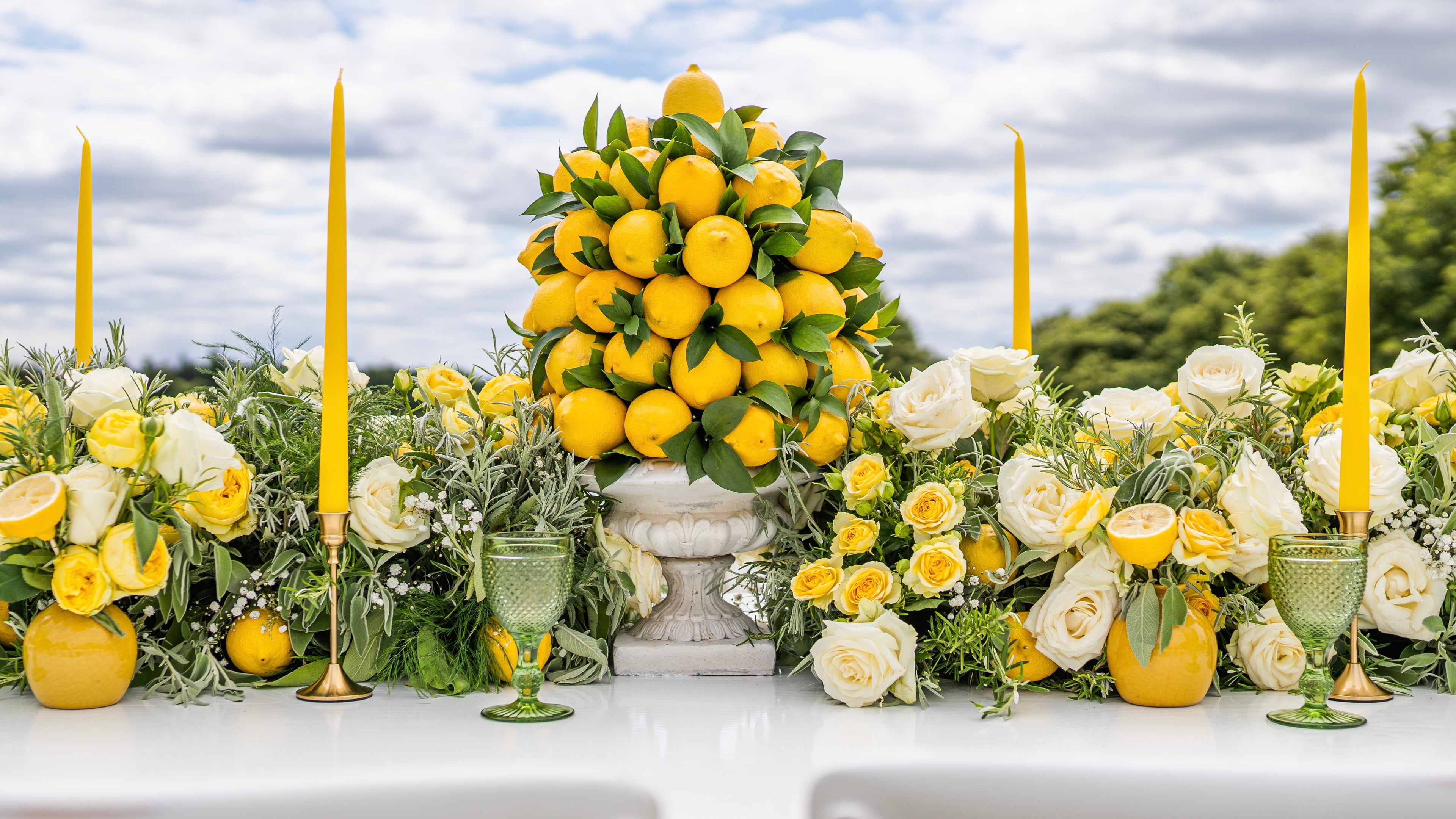 A decorative table arrangement featuring a central mound of lemons in a white pedestal bowl, flanked by white and yellow roses, green glass goblets, and tall yellow taper candles, set outdoors against a cloudy sky and trees.