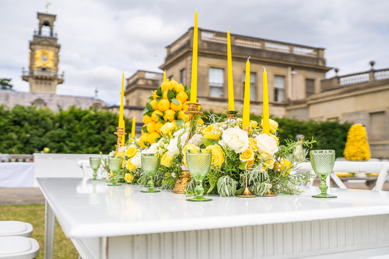 A white outdoor table set with a yellow and white floral centerpiece, tall yellow taper candles, green textured goblets, and a blurred historic building and clock tower in the background.