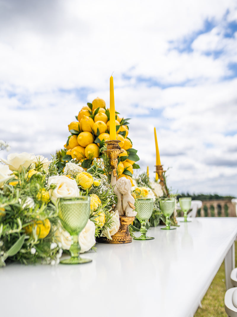 A long white table set outdoors with a decorative centerpiece featuring yellow lemons, white and yellow flowers, green glass goblets, and tall yellow taper candles under a cloudy blue sky.