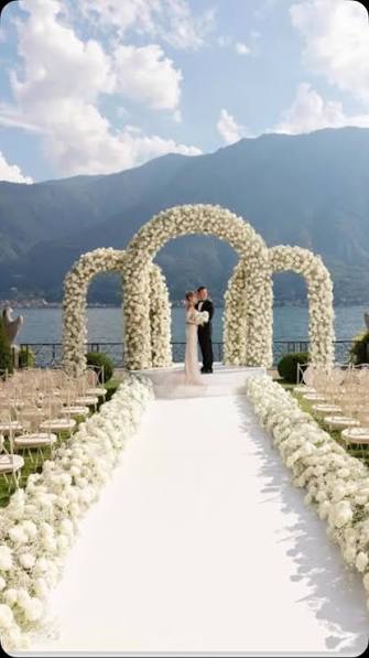 Outdoor wedding ceremony setup with a white floral archway, aisle runner, seated guests, and a couple standing under the arch with mountains and water in the background.