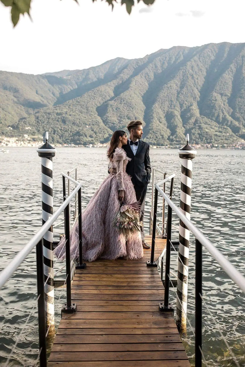 A bride in a voluminous dusty pink feathered gown and a groom in a black tuxedo stand together on a wooden dock extending over a lake, with green mountains in the background.