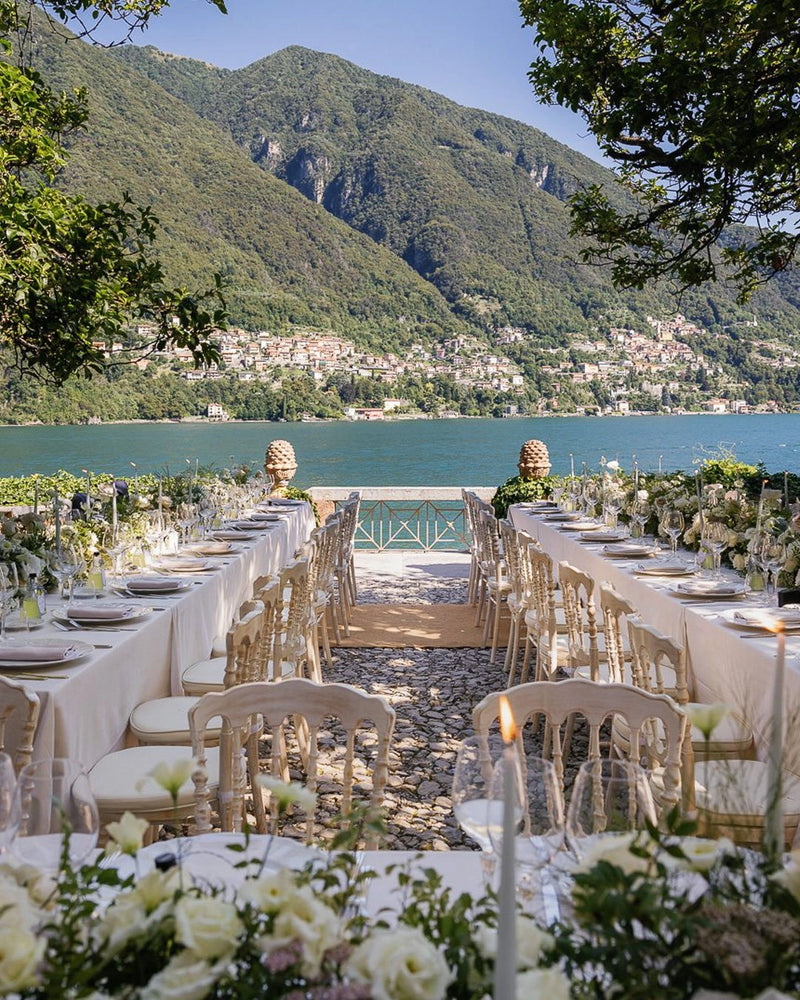 A long outdoor wedding reception table setup with white tablecloths, cream chairs, and floral centerpieces, leading toward a lakeside stage with a dramatic mountain backdrop.