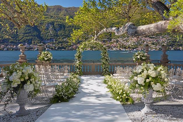 An outdoor wedding ceremony setup by a lake, featuring a white aisle runner, floral arch, white chairs, large urn-style flower arrangements, and a scenic mountain and lake background.