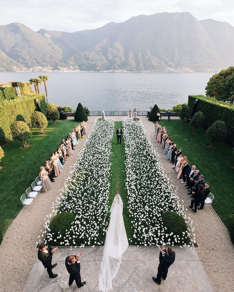 An outdoor wedding ceremony viewed from above, with a bride in a white gown standing at the altar on a stone path, flanked by guests seated on both sides. The ceremony is held in a formal garden with rows of white flowers, manicured hedges, and a large body of water with mountains in the background.