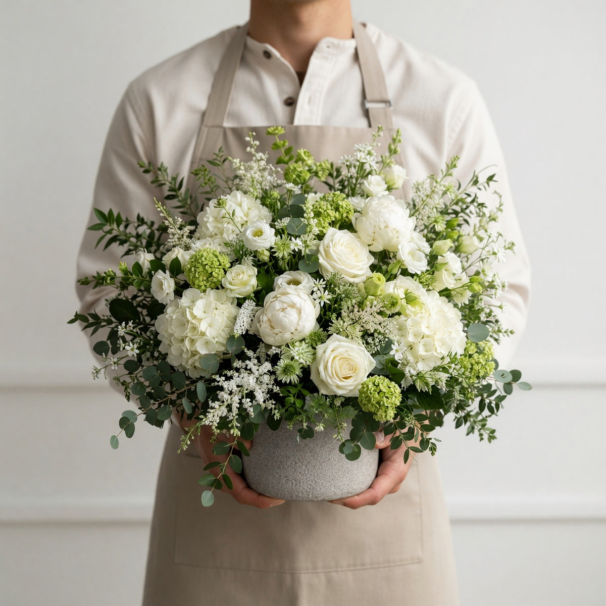 A person wearing a beige shirt and apron holds a large, lush floral arrangement in a textured gray pot. The bouquet features white roses, hydrangeas, green pompons, and eucalyptus against a clean, off-white background.