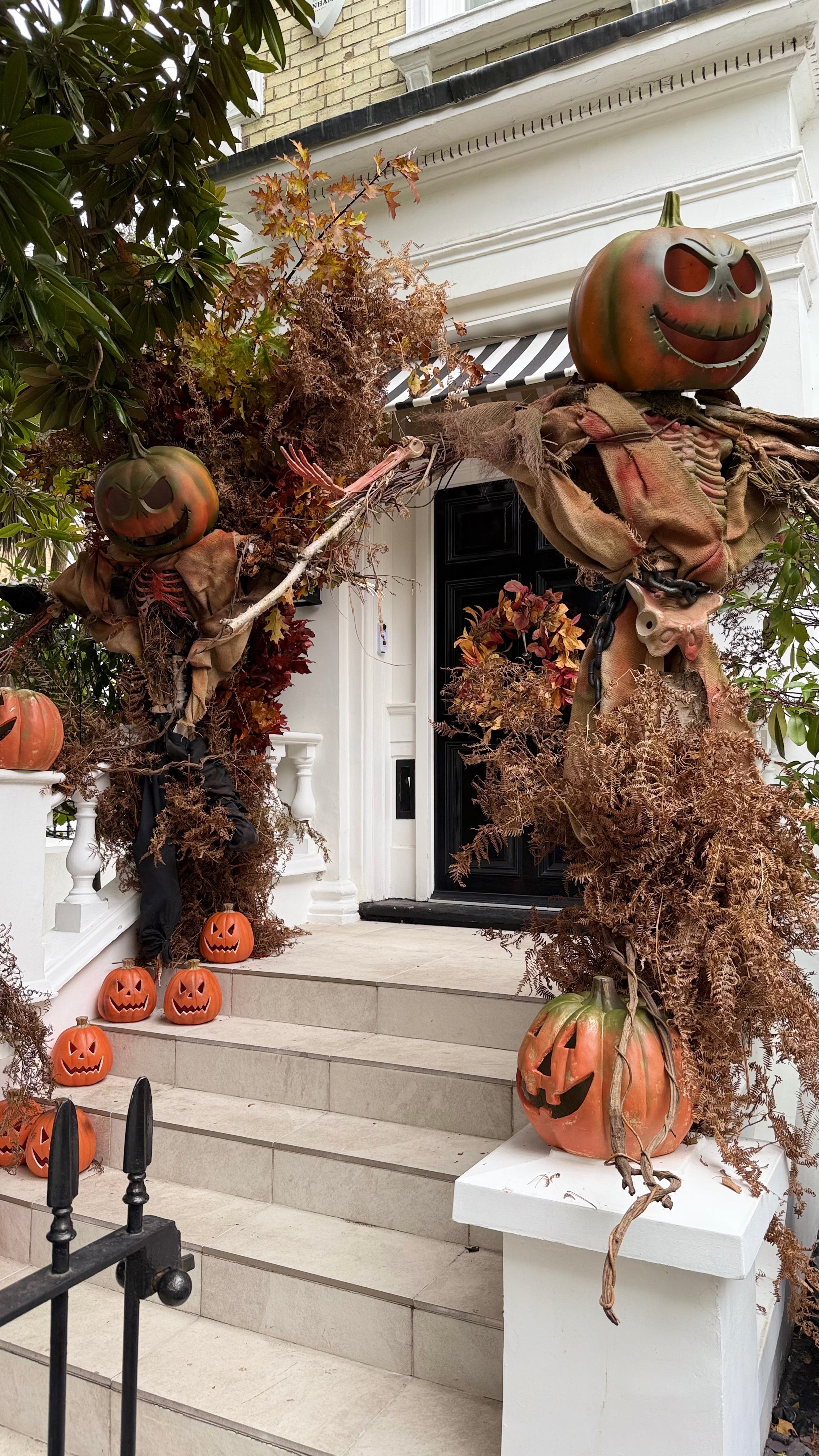 A front porch decorated for Halloween with two large scarecrows made from brown fabric and dried foliage, carved pumpkins on the steps and posts, and autumn leaves around a white door.