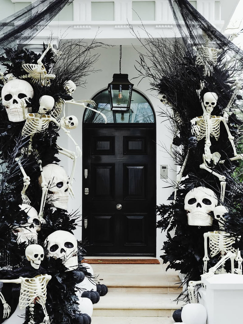 Gothic Halloween front yard decoration featuring a black door framed by black arched garlands with white skulls and skeleton figures, set against a white house facade.