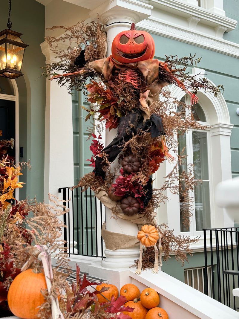A large Halloween scarecrow decoration displayed on a white porch column in front of a green house. The scarecrow features a carved orange pumpkin head, tan fabric body, black arms, and is surrounded by autumn leaves, brown branches, and small pumpkins.