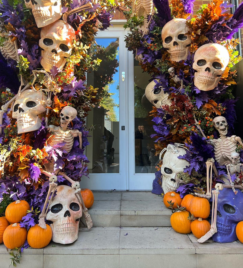 A Halloween-themed outdoor decoration setup on the steps of a building, featuring multiple decorative skulls, small skeletons, orange pumpkins, purple and autumn foliage, and a blue skull near the bottom right.