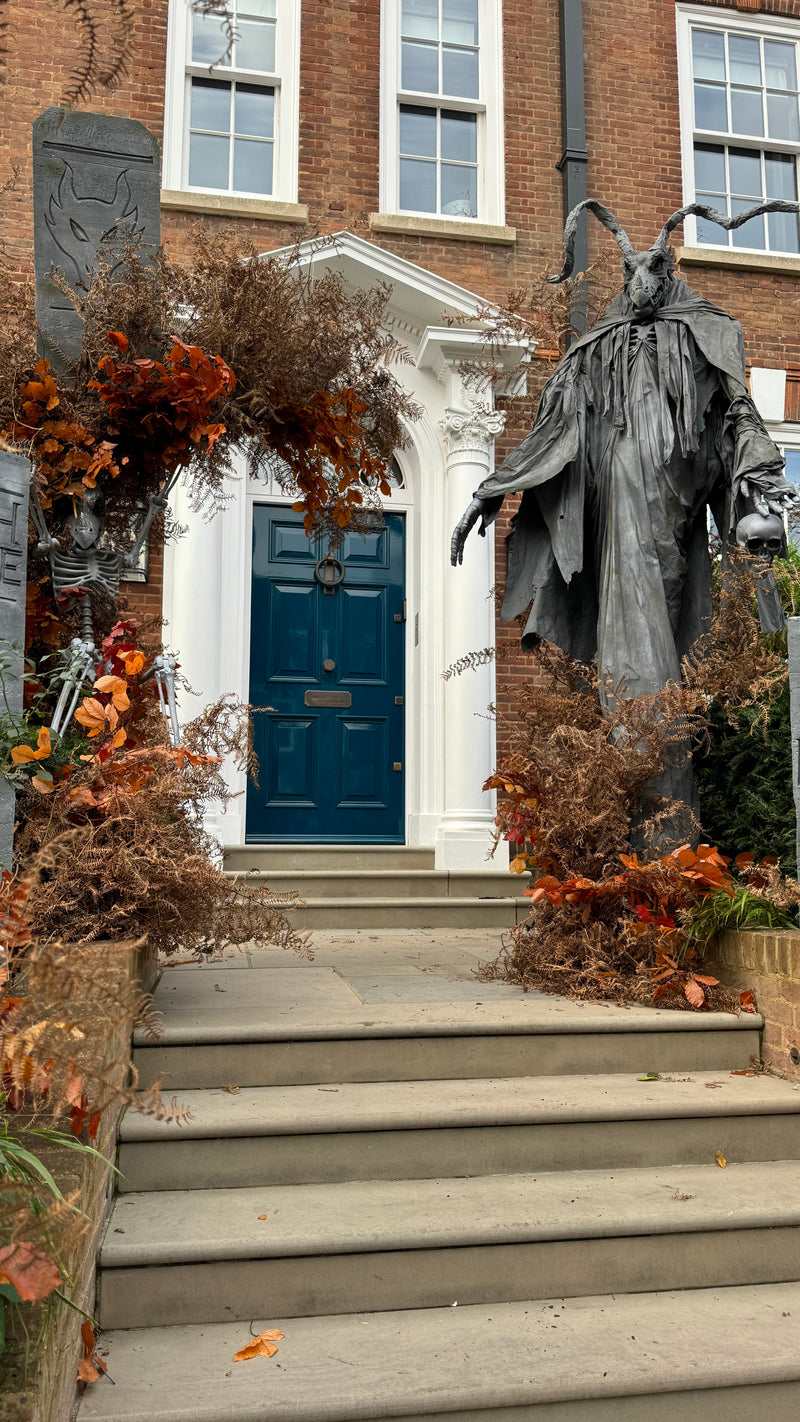 A gothic-style statue of a horned figure stands on stone steps in front of a brick building with a dark blue door, surrounded by autumn foliage and seasonal decorations.