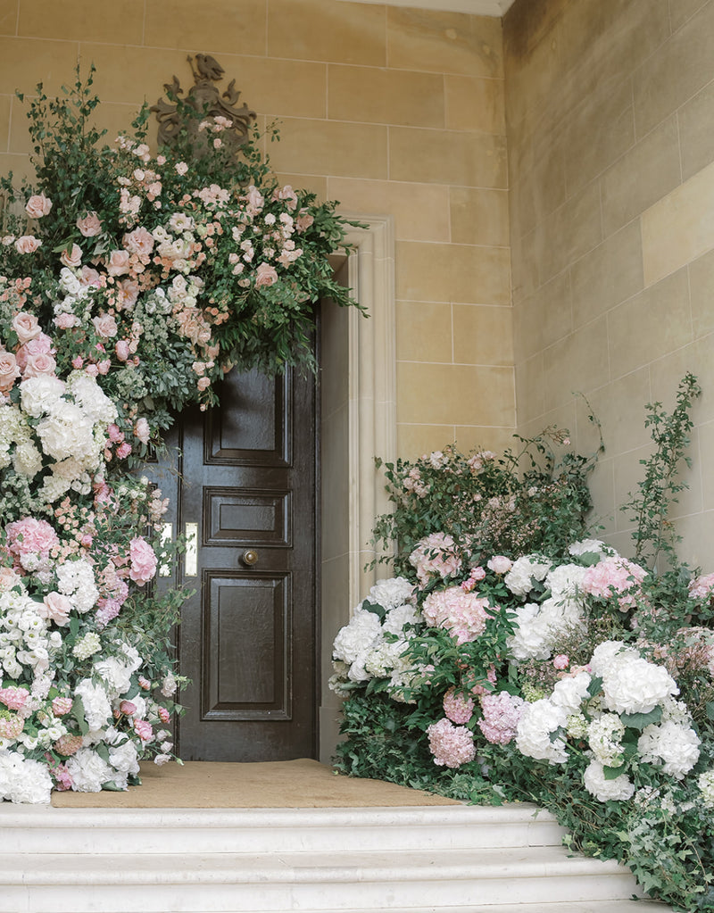 A grand dark wood door framed by lush floral arrangements in soft pink, white, and green, set against a warm beige stone wall and steps.