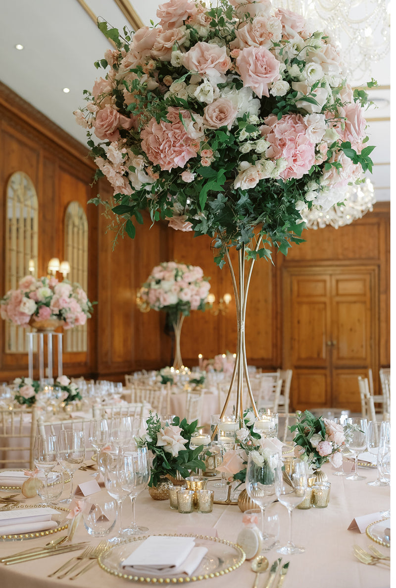 A tall floral centerpiece arrangement of blush pink and white roses with green foliage, displayed on a decorated dining table in a wood-paneled event hall.