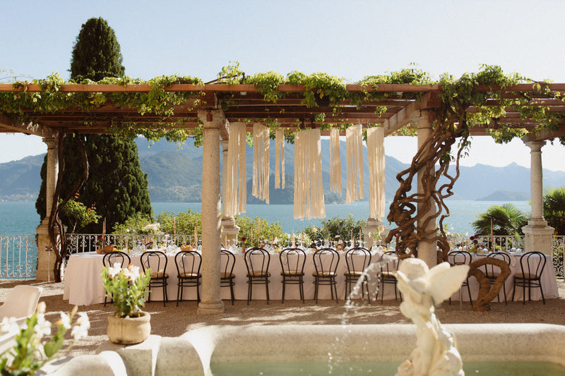 Outdoor wedding venue setup under a wooden pergola with green vines, hanging cream fabric drapes, round tables with white linens, black chairs, and a scenic lake and mountain backdrop.