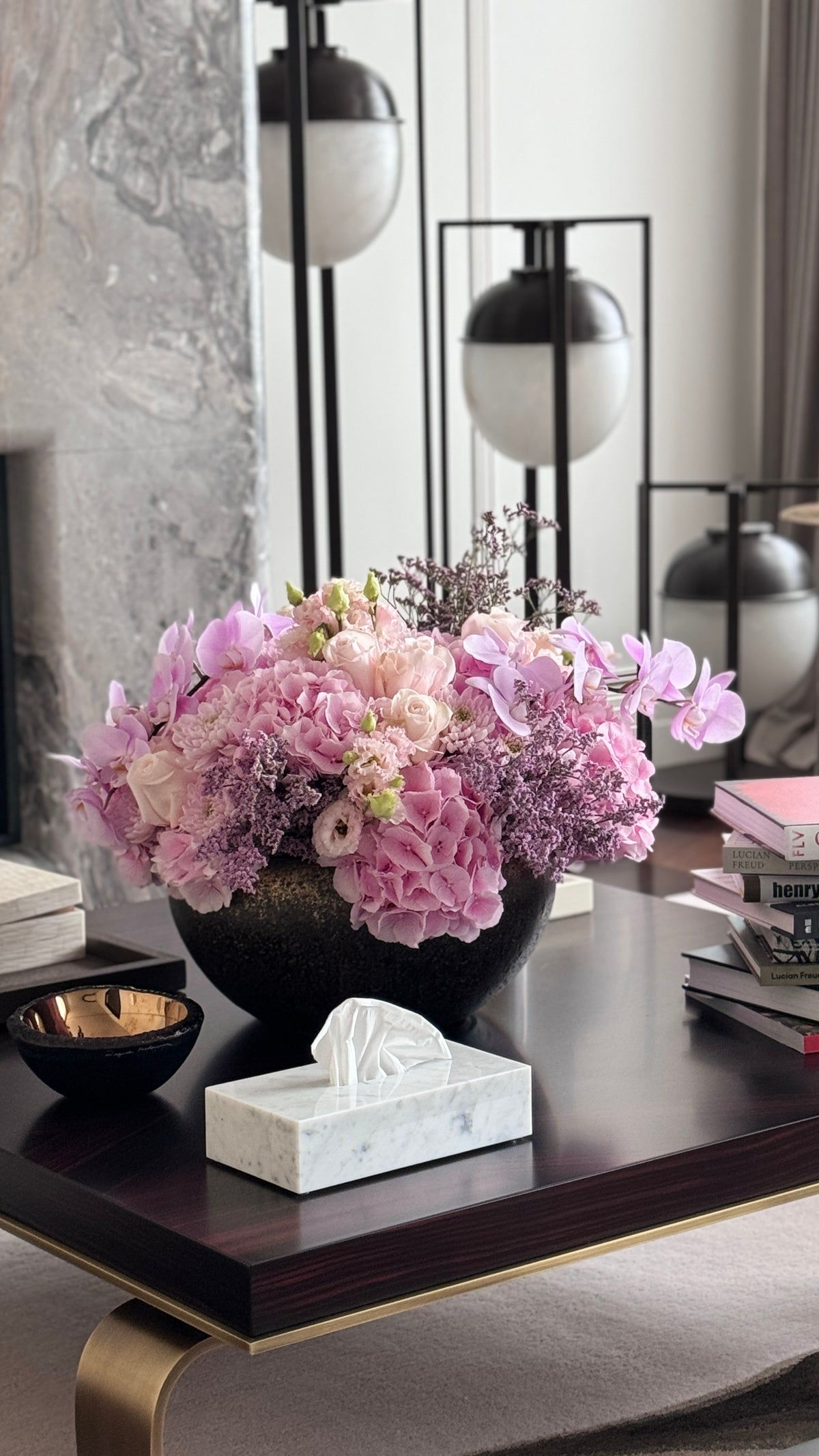 A large, vibrant floral arrangement in a dark round vase on a dark wood coffee table, styled in a modern living room setting with a marble tissue box cover, books, and contemporary lamps in the background.