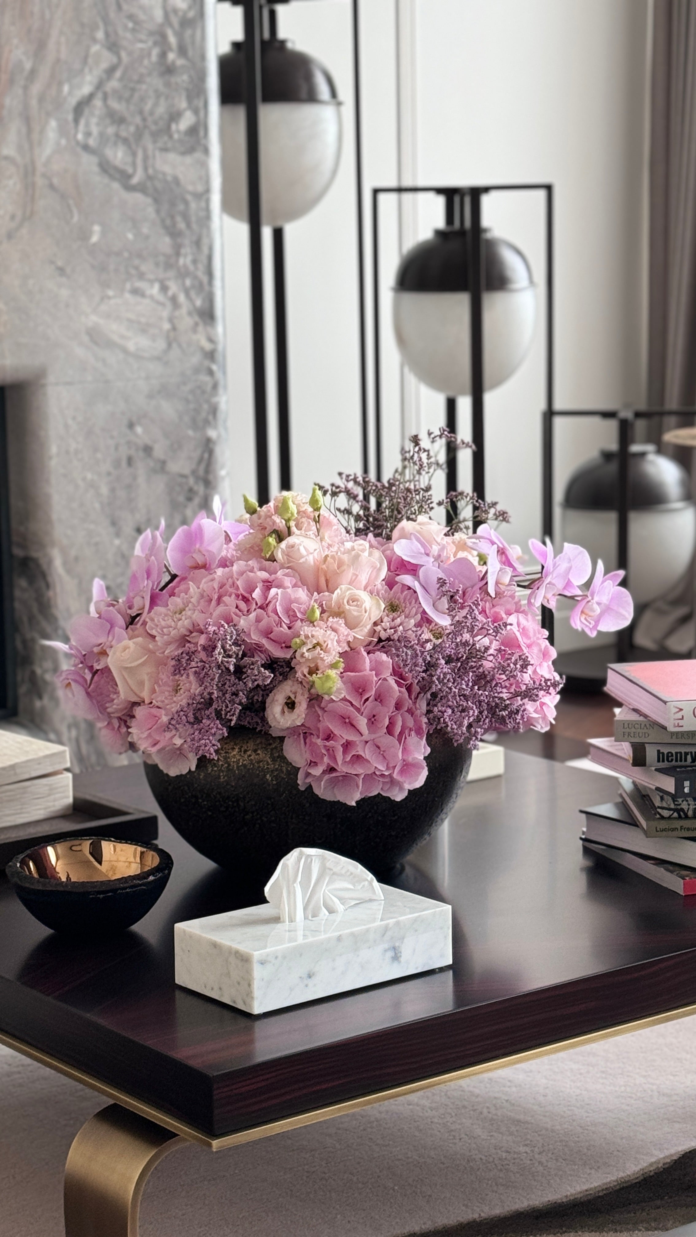 A large, vibrant floral arrangement in a dark round vase on a dark wood coffee table, styled in a modern living room setting with a marble tissue box cover, books, and contemporary lamps in the background.