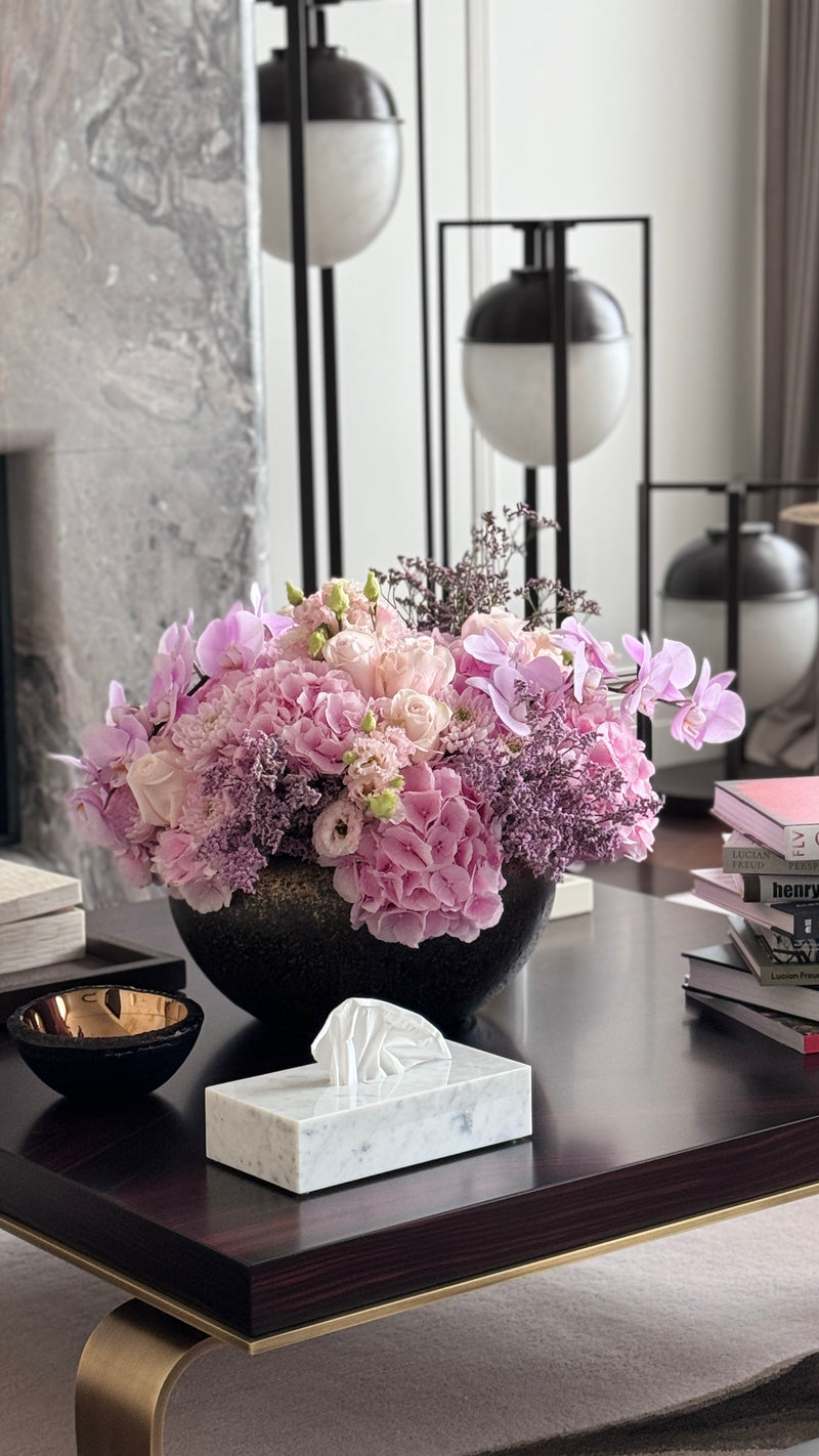 A large, vibrant floral arrangement in a dark round vase on a dark wood coffee table, styled in a modern living room setting with a marble tissue box cover, books, and contemporary lamps in the background.