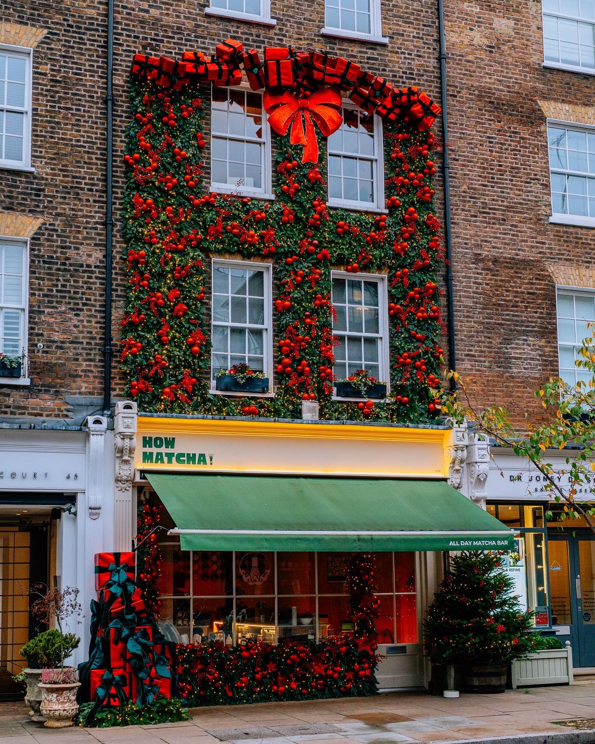 A brick building facade decorated for Christmas, with a large red bow and red floral garland covering the upper windows. The ground floor is a storefront with a green awning and a yellow sign reading 'HOW MATCHA!'.