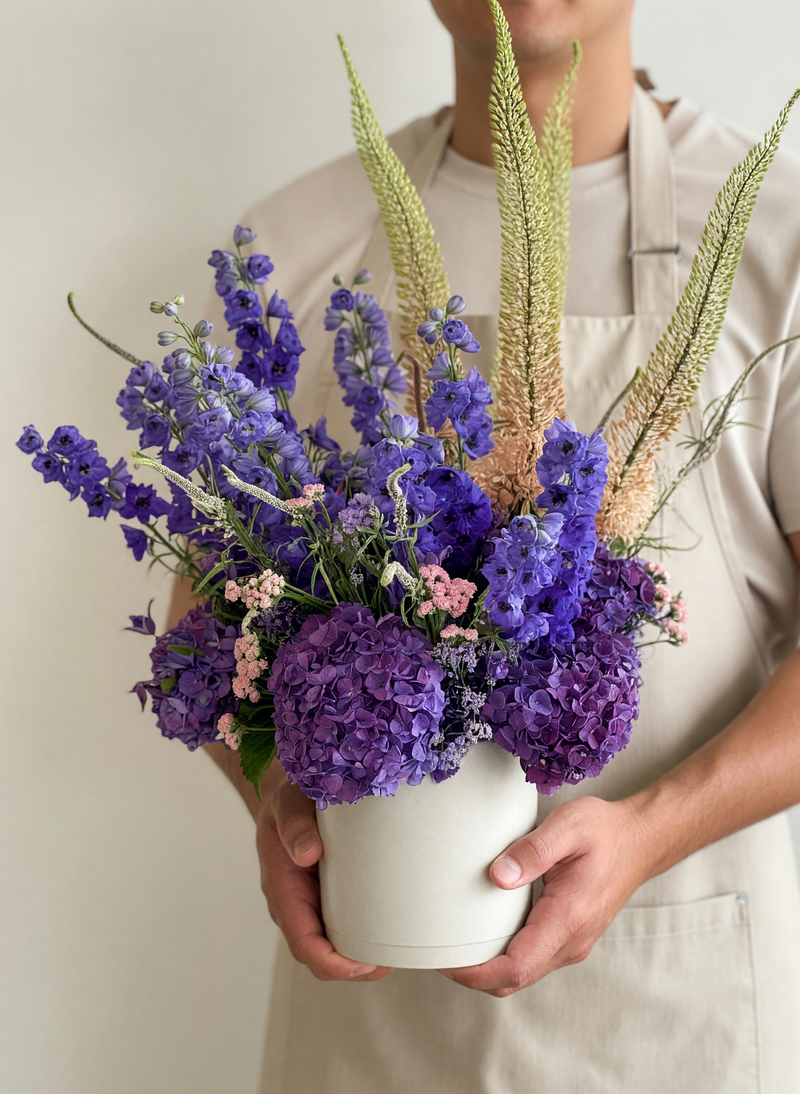 A person in a beige apron holds a white ceramic vase filled with a dried floral arrangement featuring prominent purple hydrangeas, blue delphiniums, and tan ornamental grasses against a plain light background.