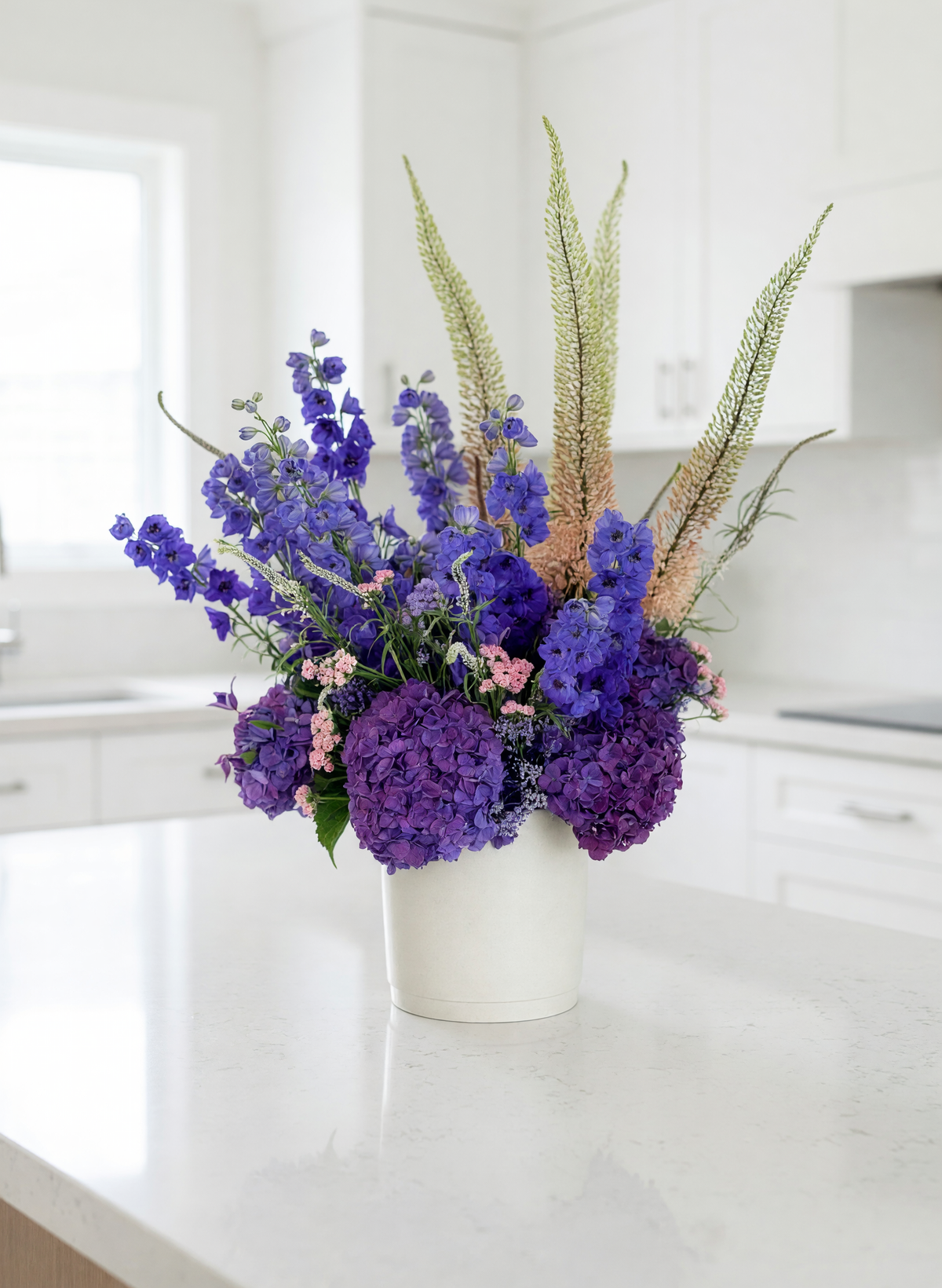 A vibrant floral arrangement featuring purple hydrangeas, blue delphiniums, and beige ornamental grasses in a white ceramic pot, displayed on a white kitchen countertop.