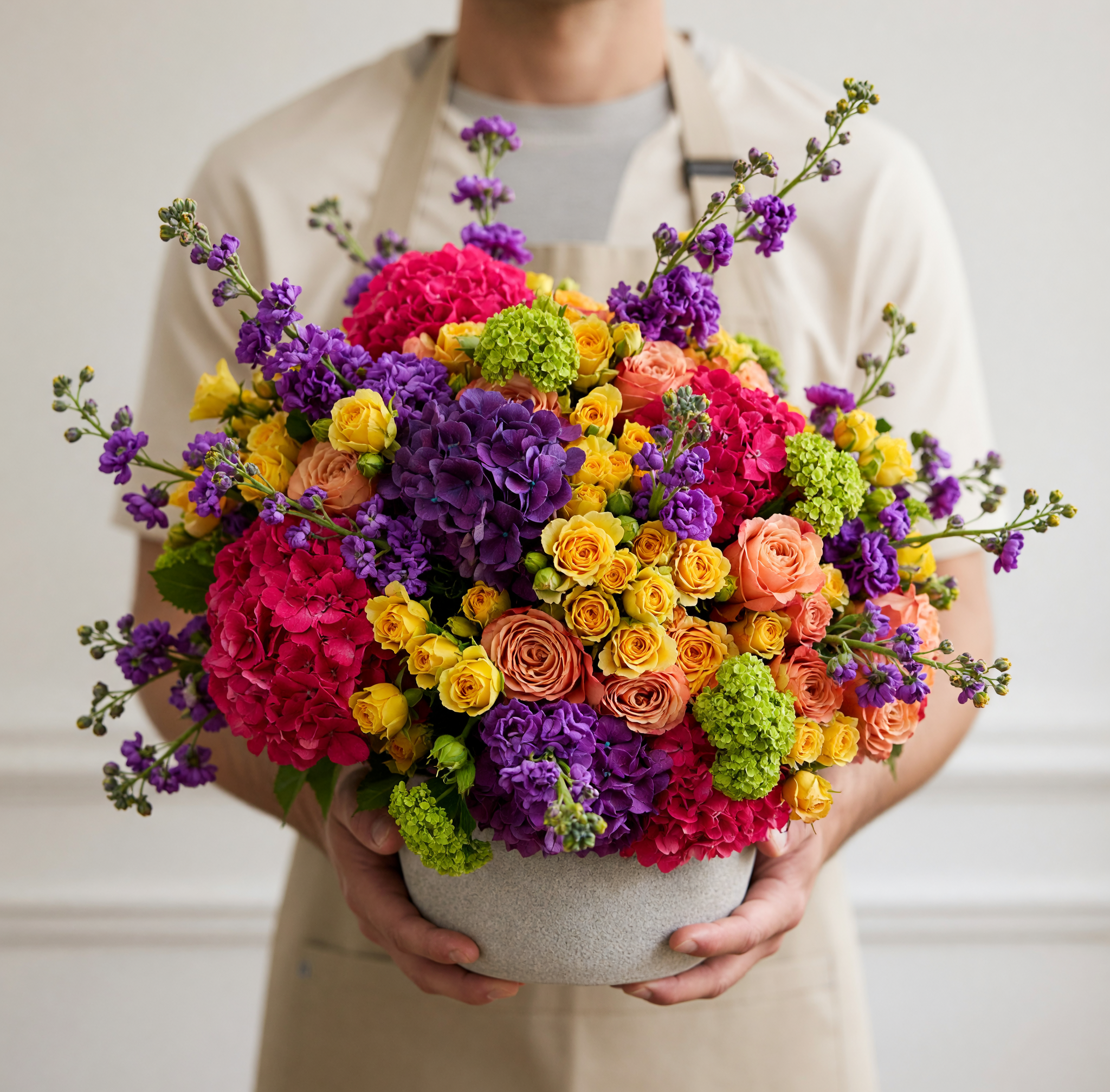 A person in a beige apron holds a large, vibrant flower bouquet in a gray ceramic pot. The bouquet features a colorful mix of pink, purple, yellow, orange, and green flowers.