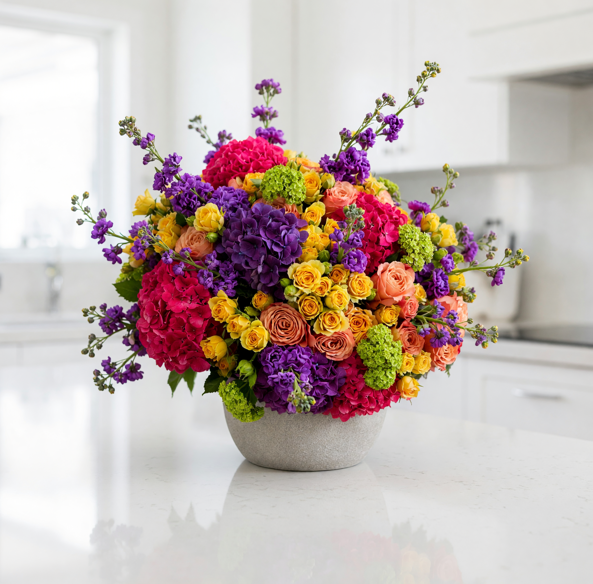 A vibrant, multicolored floral arrangement featuring pink, purple, yellow, and orange flowers in a textured gray ceramic pot, set on a white kitchen countertop with a blurred kitchen background.