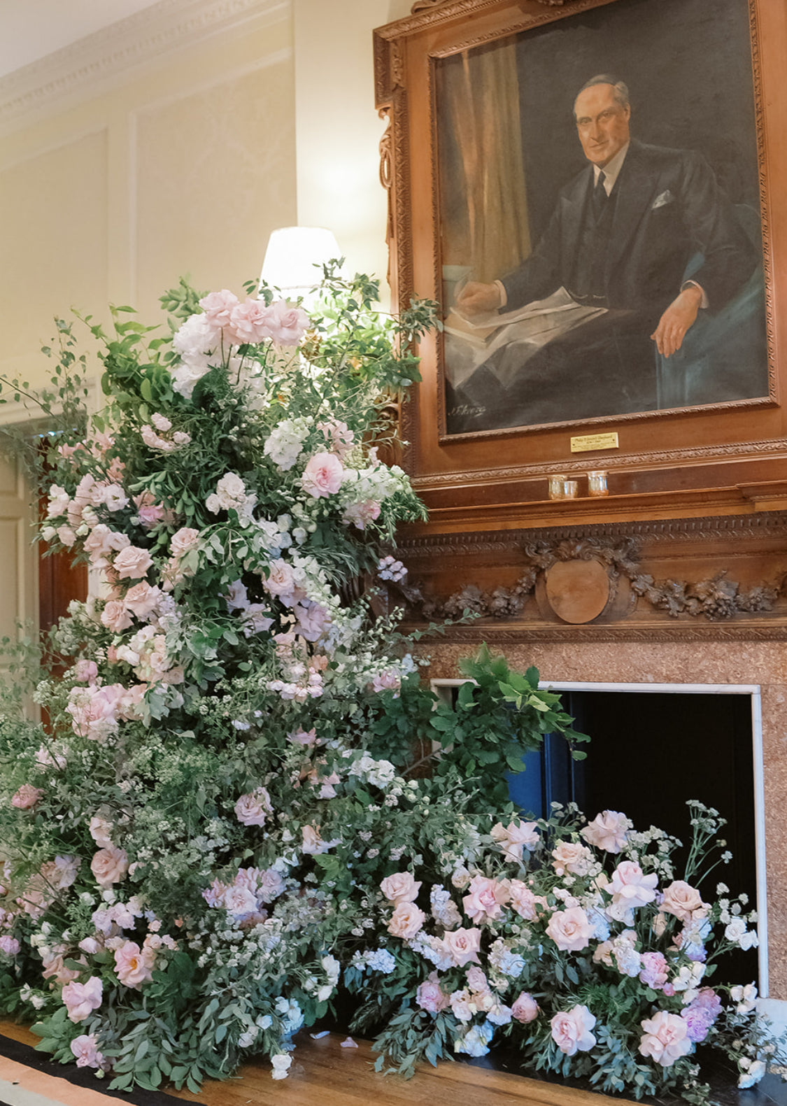 A large floral arrangement in soft pink and white roses with green foliage, displayed on a fireplace mantel beneath a framed portrait of a man in a suit.