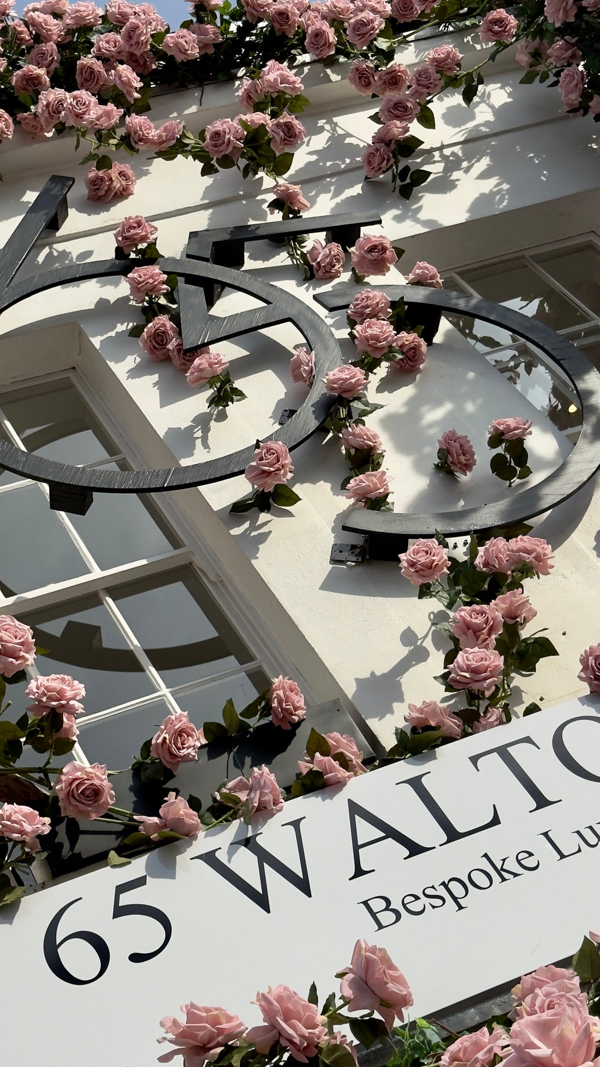 A white building facade decorated with numerous pink roses and green leaves, featuring a large black bicycle emblem and a white sign that reads '65 WALTO Bespoke Luxe'.