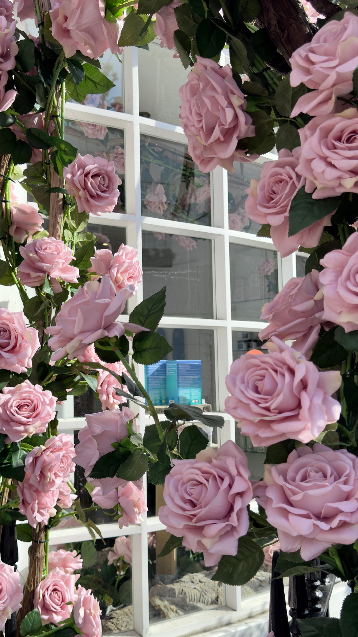 Pink artificial roses with green leaves draped over a white-framed window, creating a floral arch and backdrop.