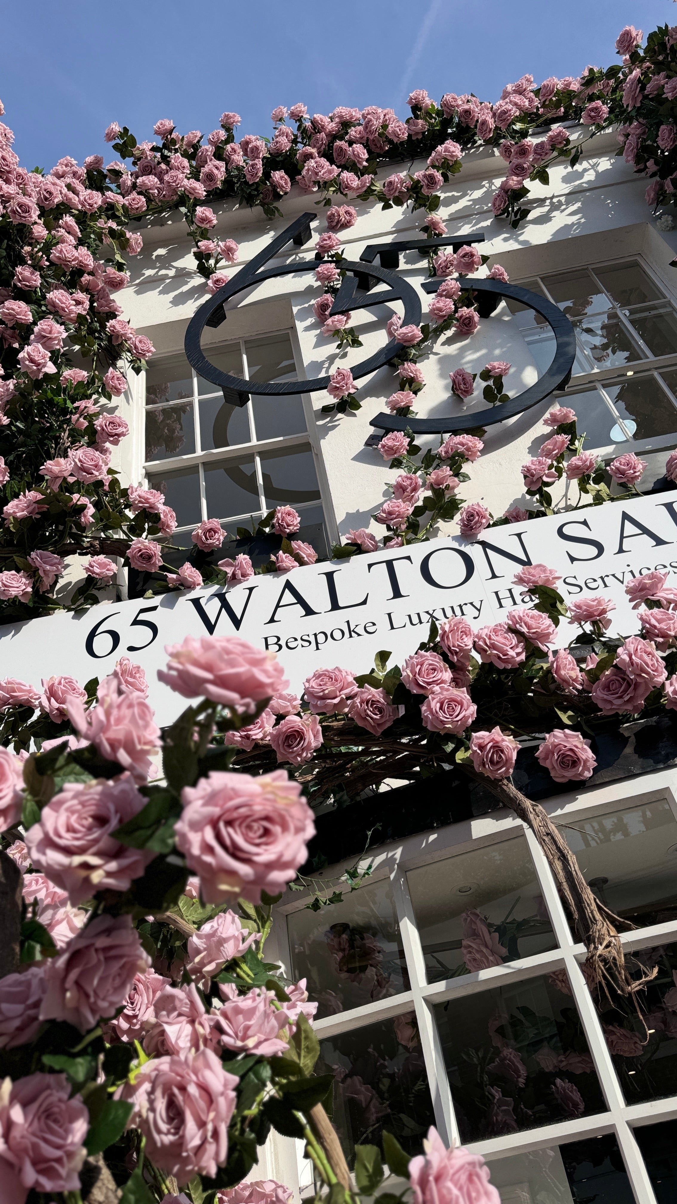 A white building facade covered with abundant pink roses, featuring a large black decorative '65' sign and a white sign reading '65 WALTON S.