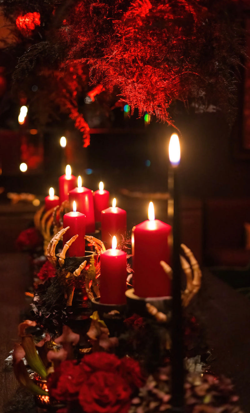 A moody arrangement of lit red pillar candles in a gothic setting, surrounded by dark foliage, red roses, and skeletal hand props under dramatic red lighting.