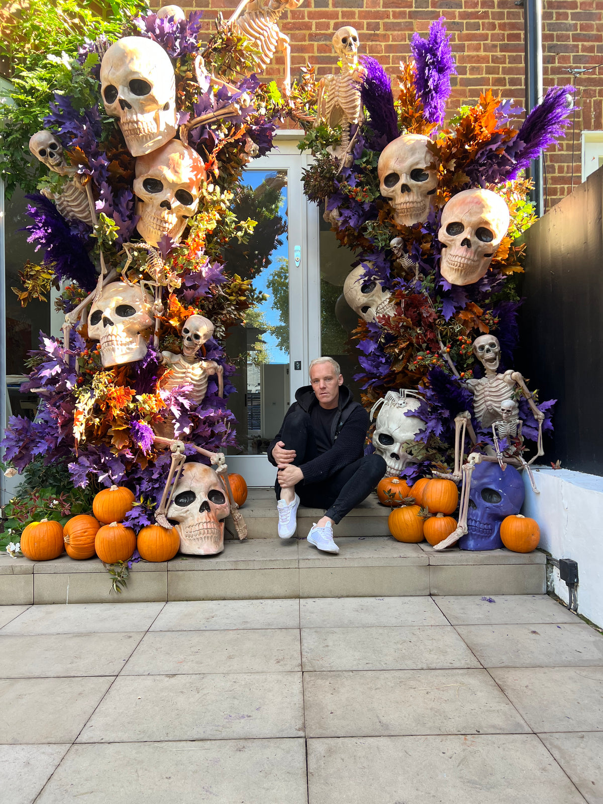 A man sits on steps in front of a building entrance decorated with large Halloween skeletons, skulls, purple feathers, orange pumpkins, and autumn foliage.