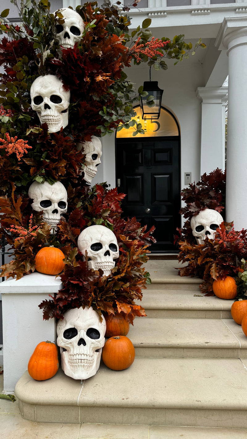 Halloween front porch decorated with large white skulls, orange pumpkins, and autumn leaves draped over stone steps and a black door.