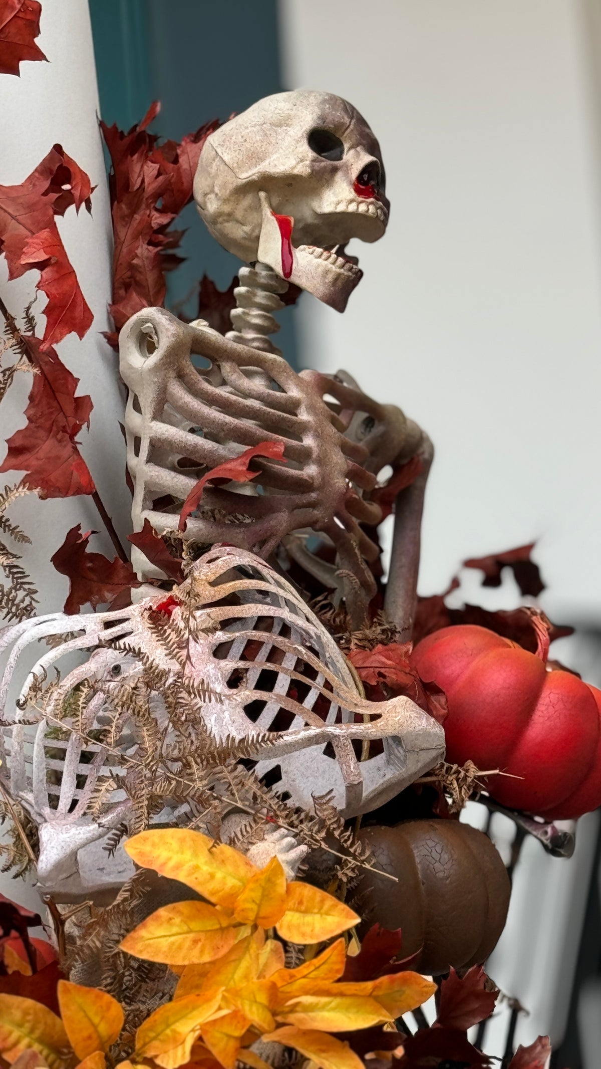 A Halloween decorative skeleton figurine posed among autumn leaves, pumpkins, and ferns, with a red apple near its skull.