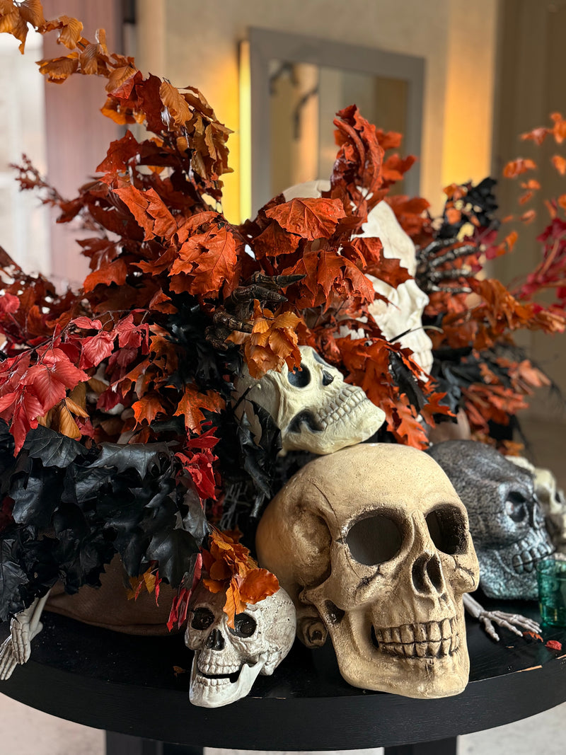 Halloween decorative centerpiece featuring multiple realistic skulls in cream, gray, and black, surrounded by red, orange, and black faux autumn leaves on a black table.