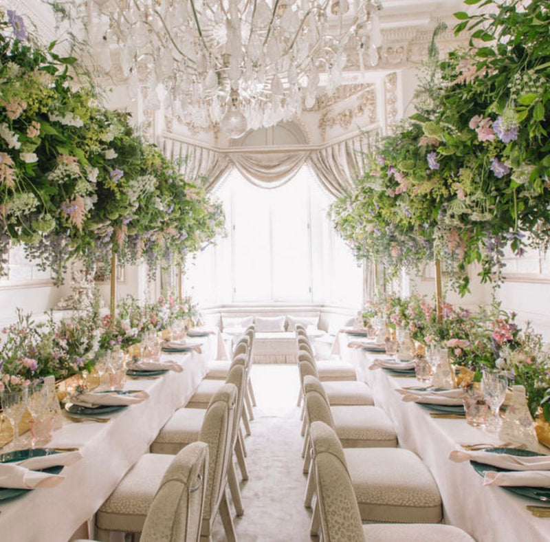 A grand, elegant event space with long dining tables set with white linens, floral centerpieces, and chairs, under a large crystal chandelier and draped windows.