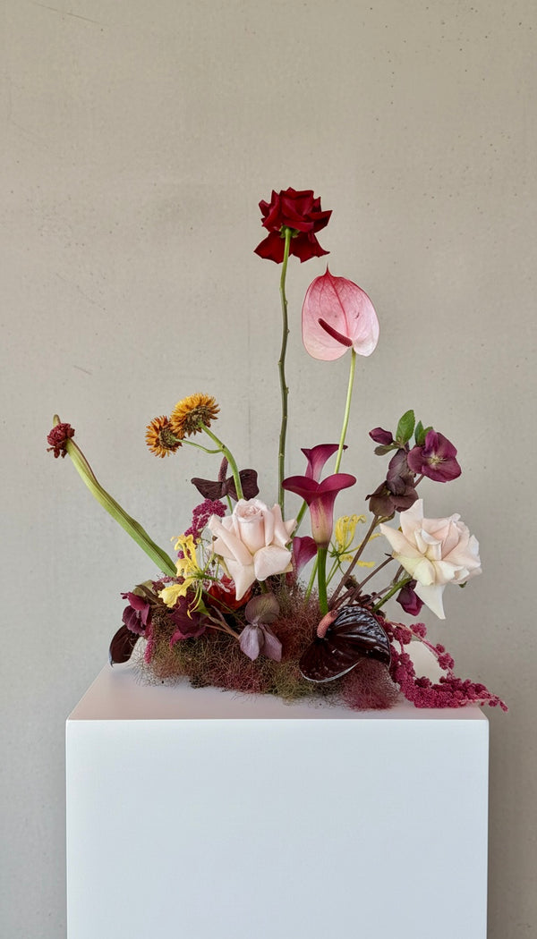 A floral arrangement featuring a dark red rose, a pink anthurium, pale blush roses, purple orchids, and small yellow flowers, displayed on a white pedestal against a neutral beige wall.