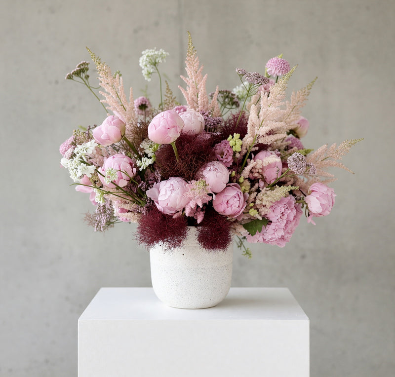 A floral arrangement of pink peonies, dusty pink ornamental grasses, burgundy filler flowers, and small white blossoms in a textured white ceramic vase, displayed on a white pedestal against a gray concrete wall.